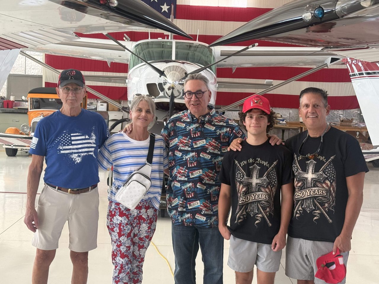 Five guests in flag shirts and 250 years tees under a plane wing in the hangar