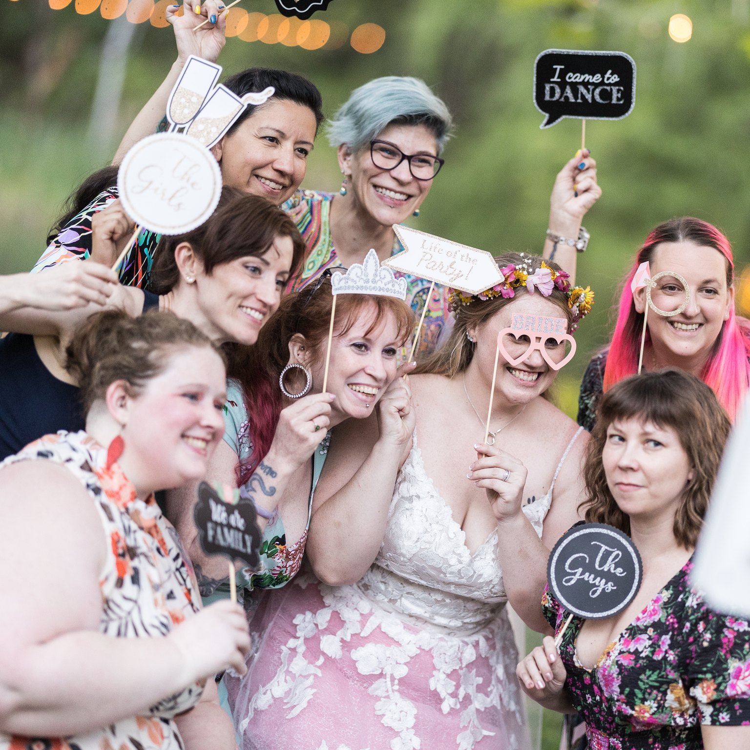 Abney and her wedding guests laugh behind photo booth props, holding an I came to dance sign and heart shaped glasses