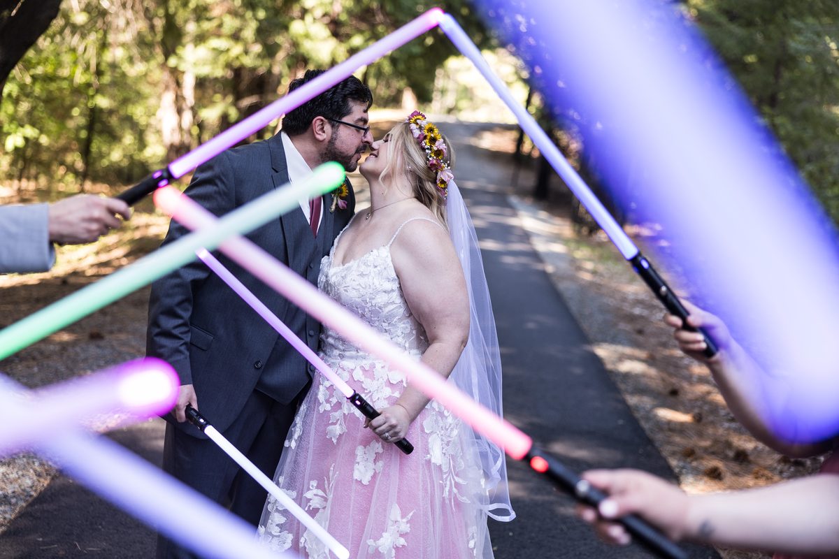 Chris kisses Abney on a forest path, framed by glowing purple, pink and green lightsabers held by the wedding party