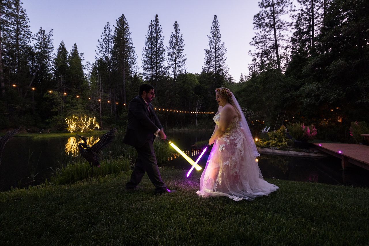 Chris holds a yellow lightsaber and Abney a purple one on the lawn by the pond