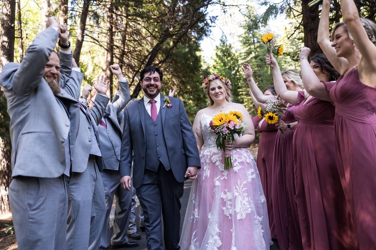 Abney and Chris look at the camera while their wedding party cheers around them on a forest path