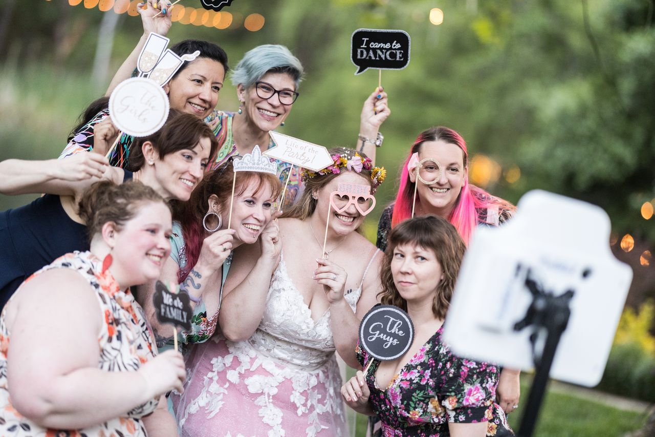 Abney and her wedding guests laugh behind photo booth props, holding an I came to dance sign and heart shaped glasses