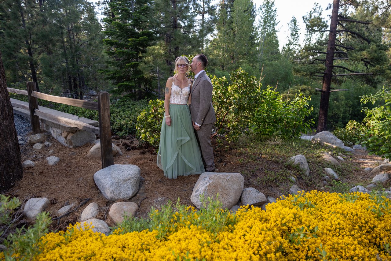 Amy and Justin stand on the hillside by a wooden bench and wildflowers