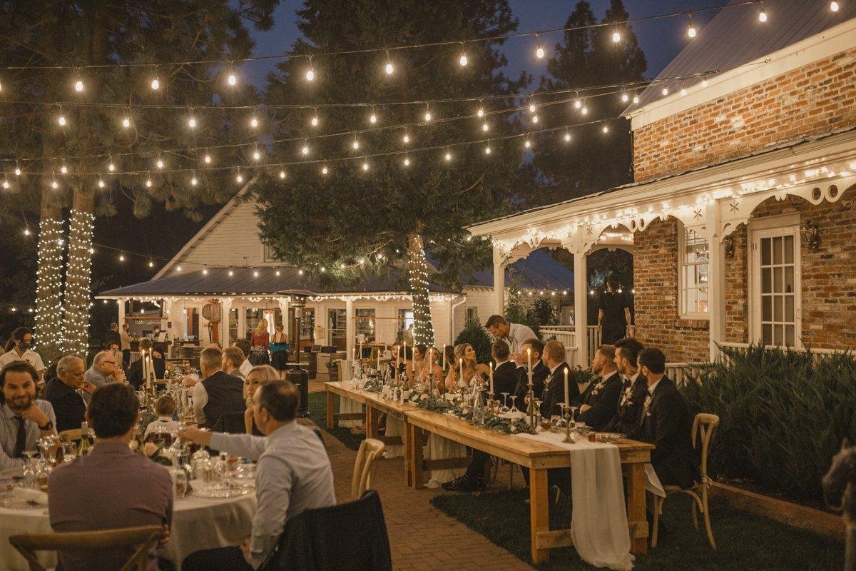 Long tables of guests at dinner beside the brick house under string lights, photo by Composing Reality Photography