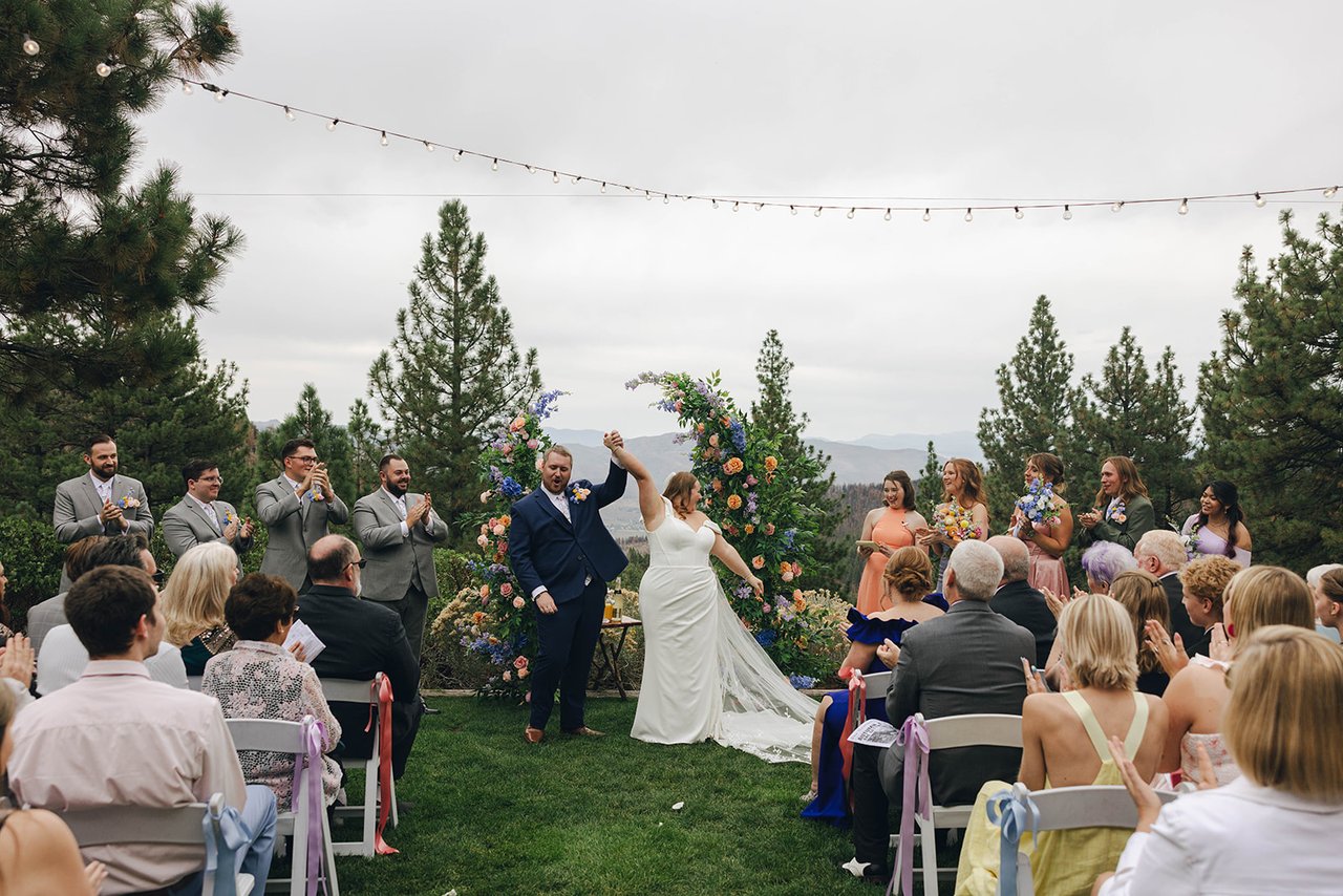 The newlyweds celebrate at the floral arch with an arm thrown in the air while guests clap