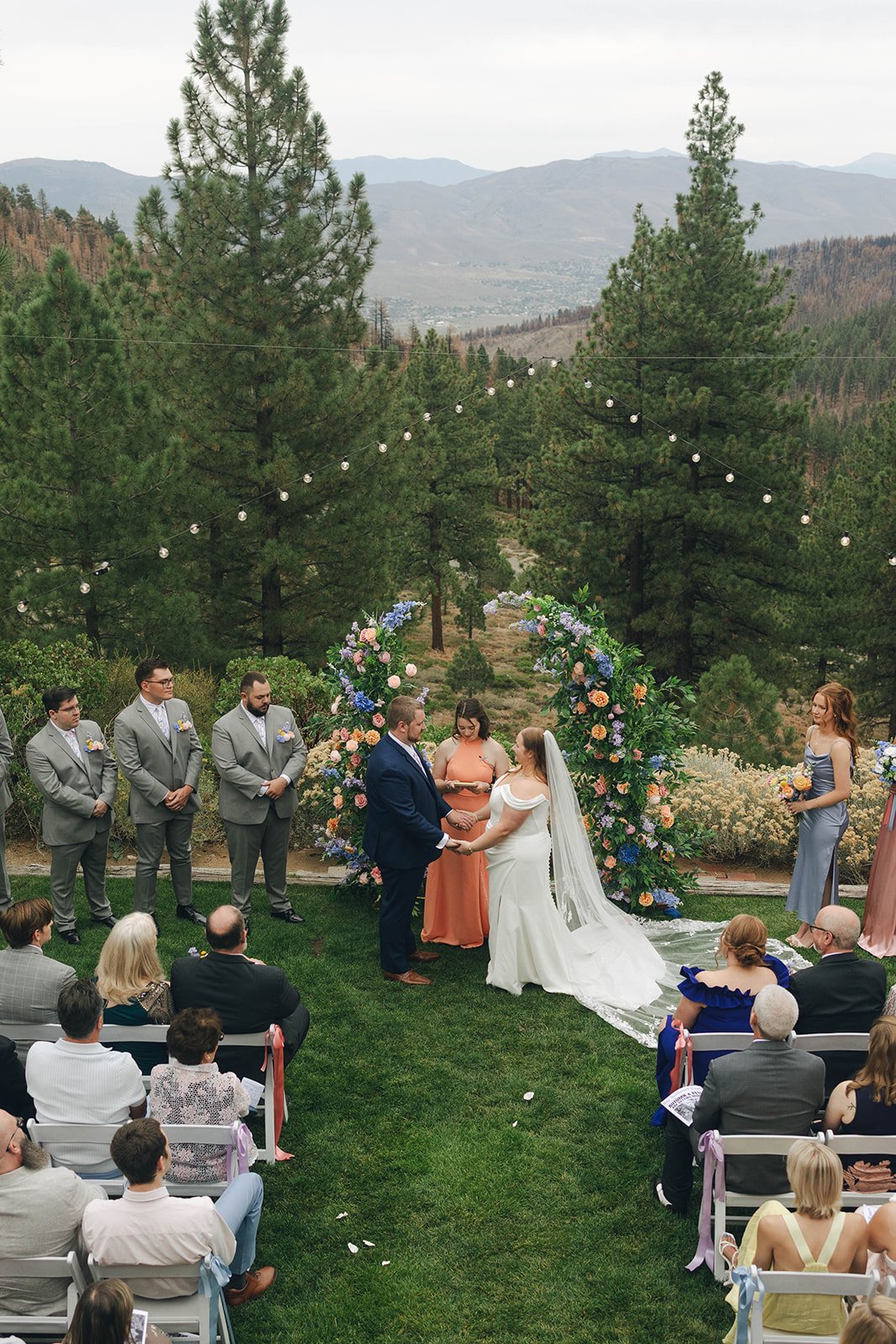 Autumn and Peter hold hands at the floral arch, seen from above, with guests seated on the lawn and the mountains beyond