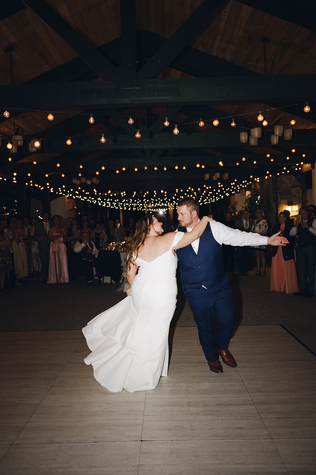 Autumn and Peter dance with arms out during their first dance, her dress swirling under the string lights
