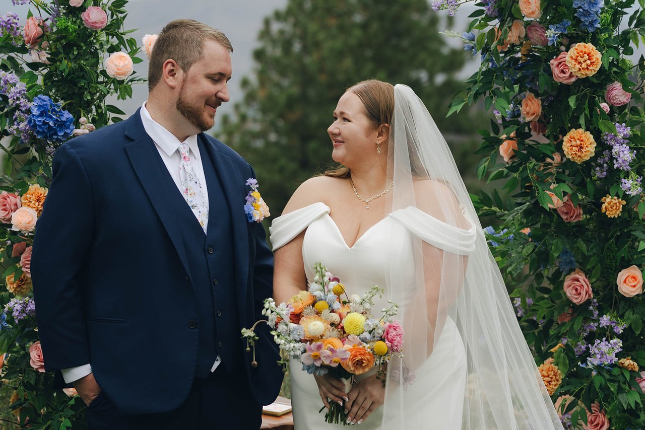 Autumn and Peter smile at each other between the two sides of the floral arch