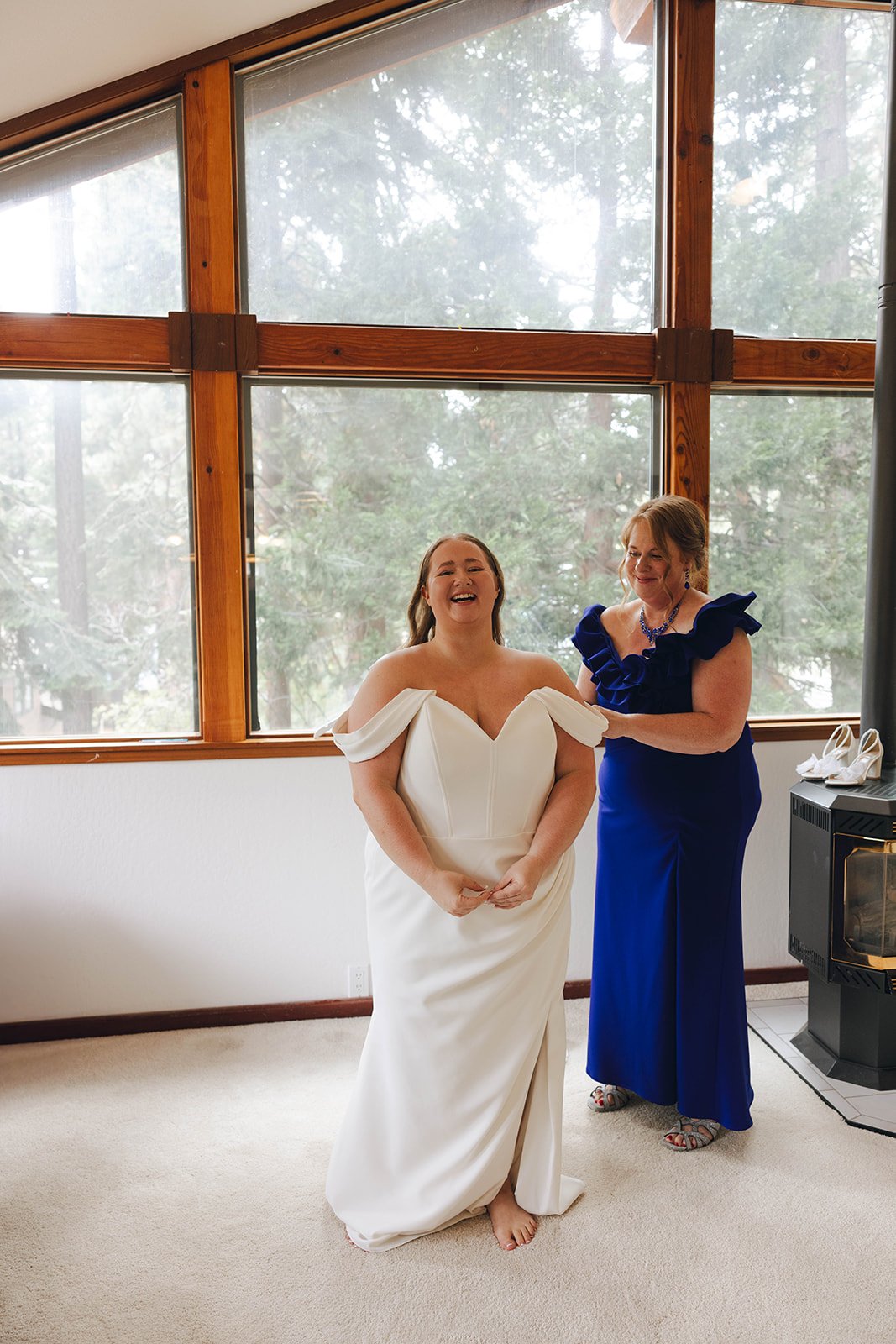 Autumn laughs in her wedding dress as it is fastened, with tall windows and pines behind her