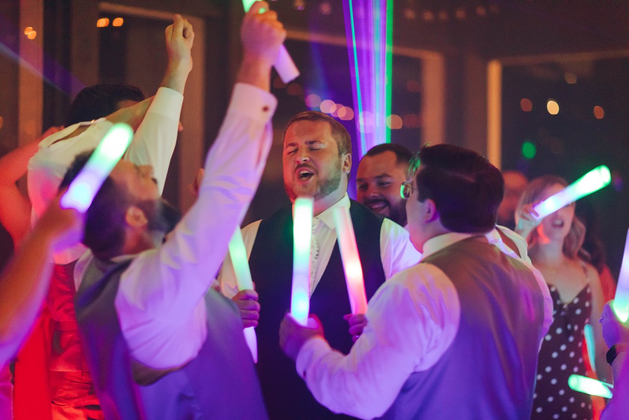 Groomsmen sing with glow sticks raised under magenta and green laser beams