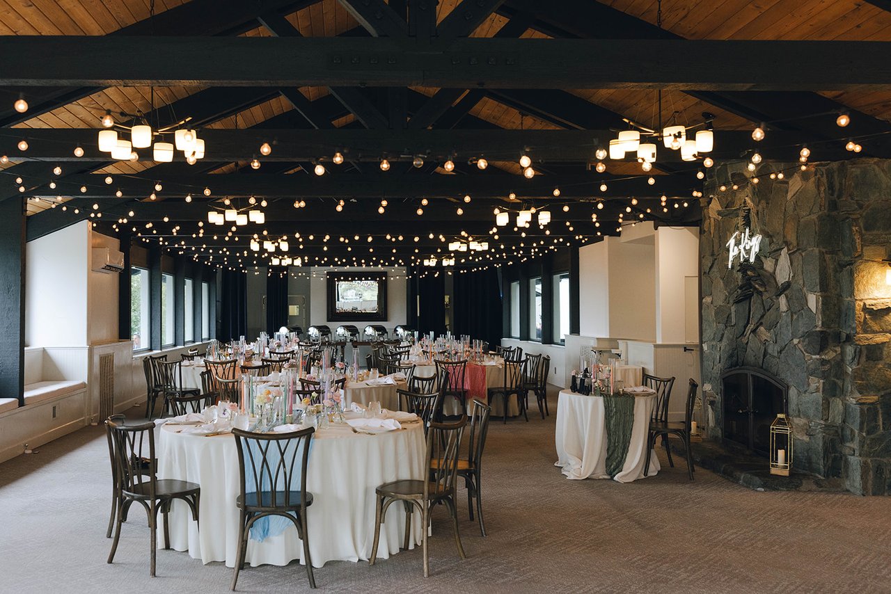 The reception room at the Tannenbaum Event Center set for dinner, with string lights under the wood beam ceiling and a stone fireplace