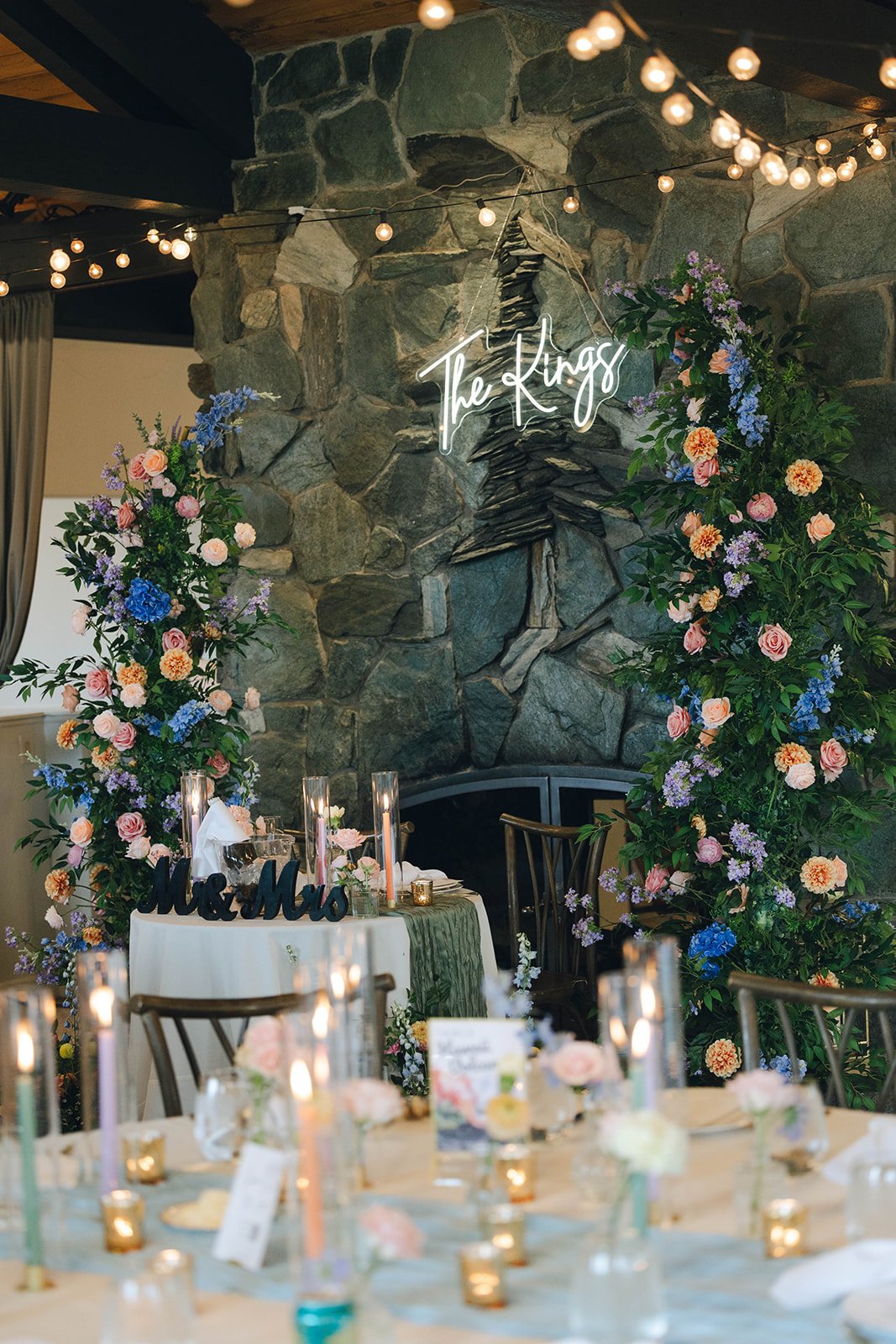 The sweetheart table at the stone fireplace, framed by flowers and a neon sign reading The Kings