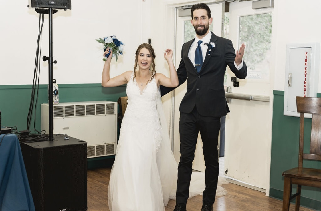 Caitlyn raises her blue and white bouquet and John throws his arms wide as they make their grand entrance