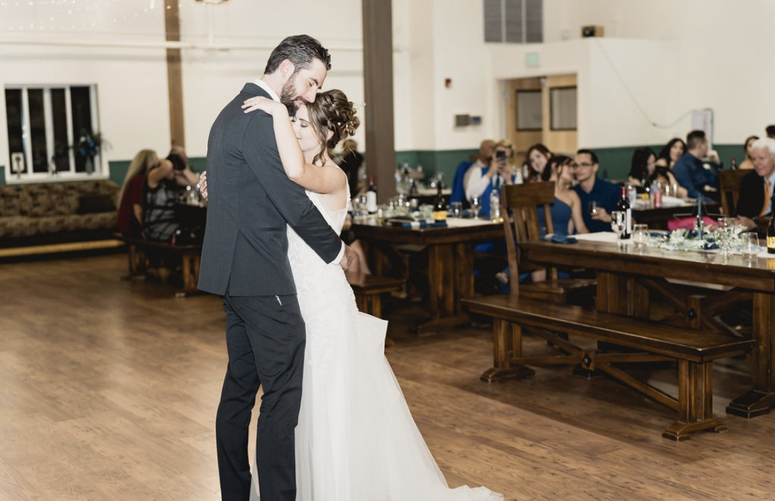 Caitlyn and John share a slow dance on the wood floor while their guests watch from the long tables