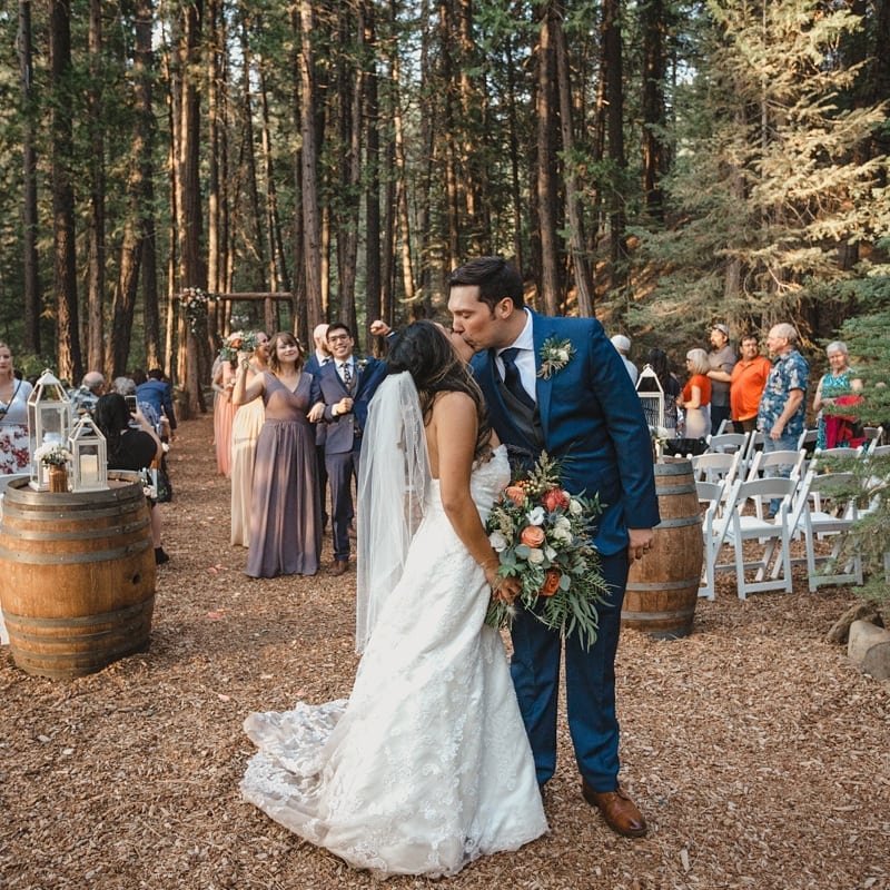 Hannah and Kyle kiss in the forest aisle between wine barrels and lanterns as their bridesmaids cheer