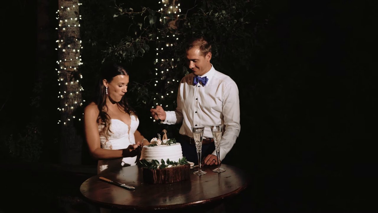 Jen and Wes cut their white wedding cake at a small table under the string lights