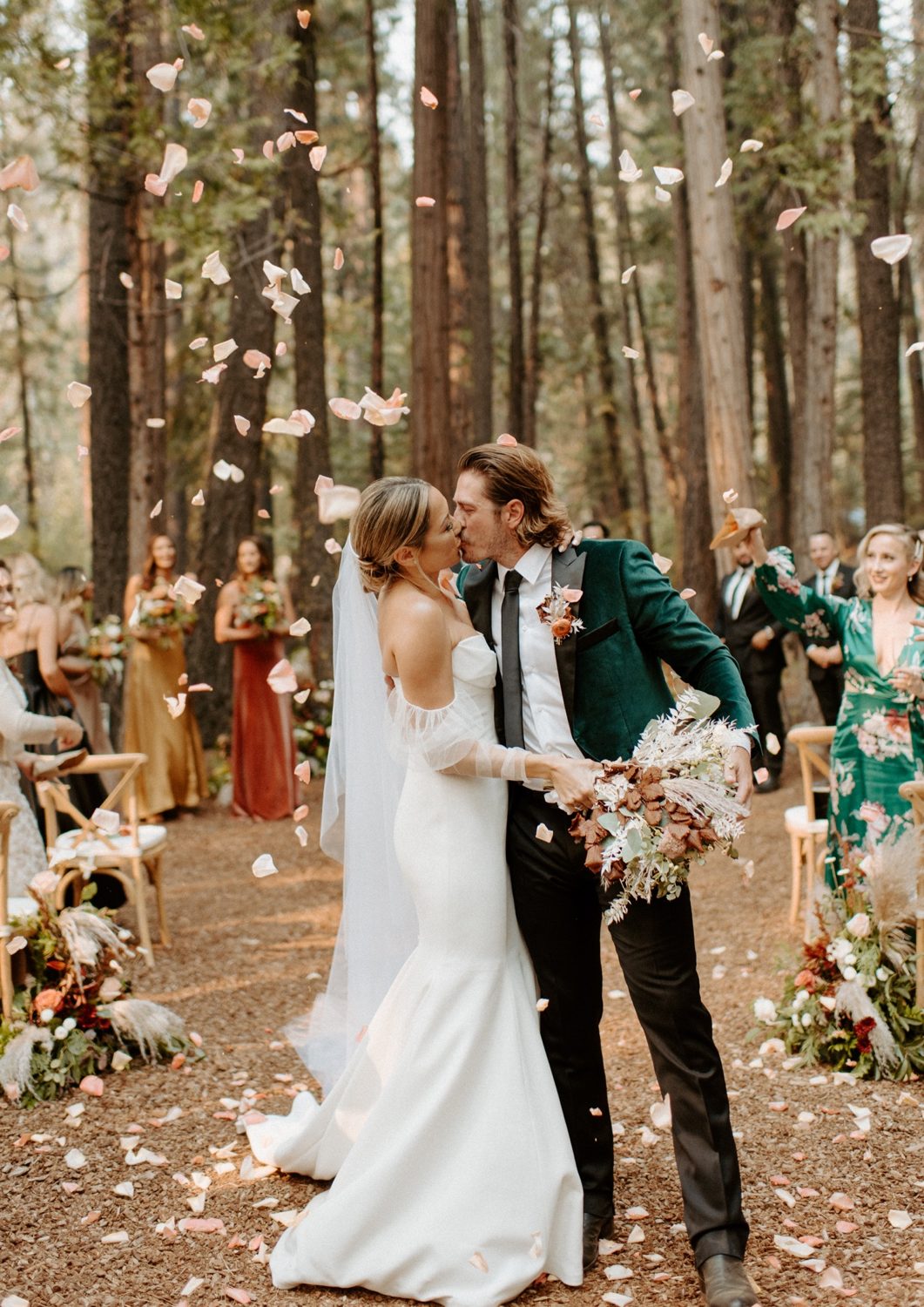 Joan and Kyle kiss as flower petals fly at the end of their forest ceremony