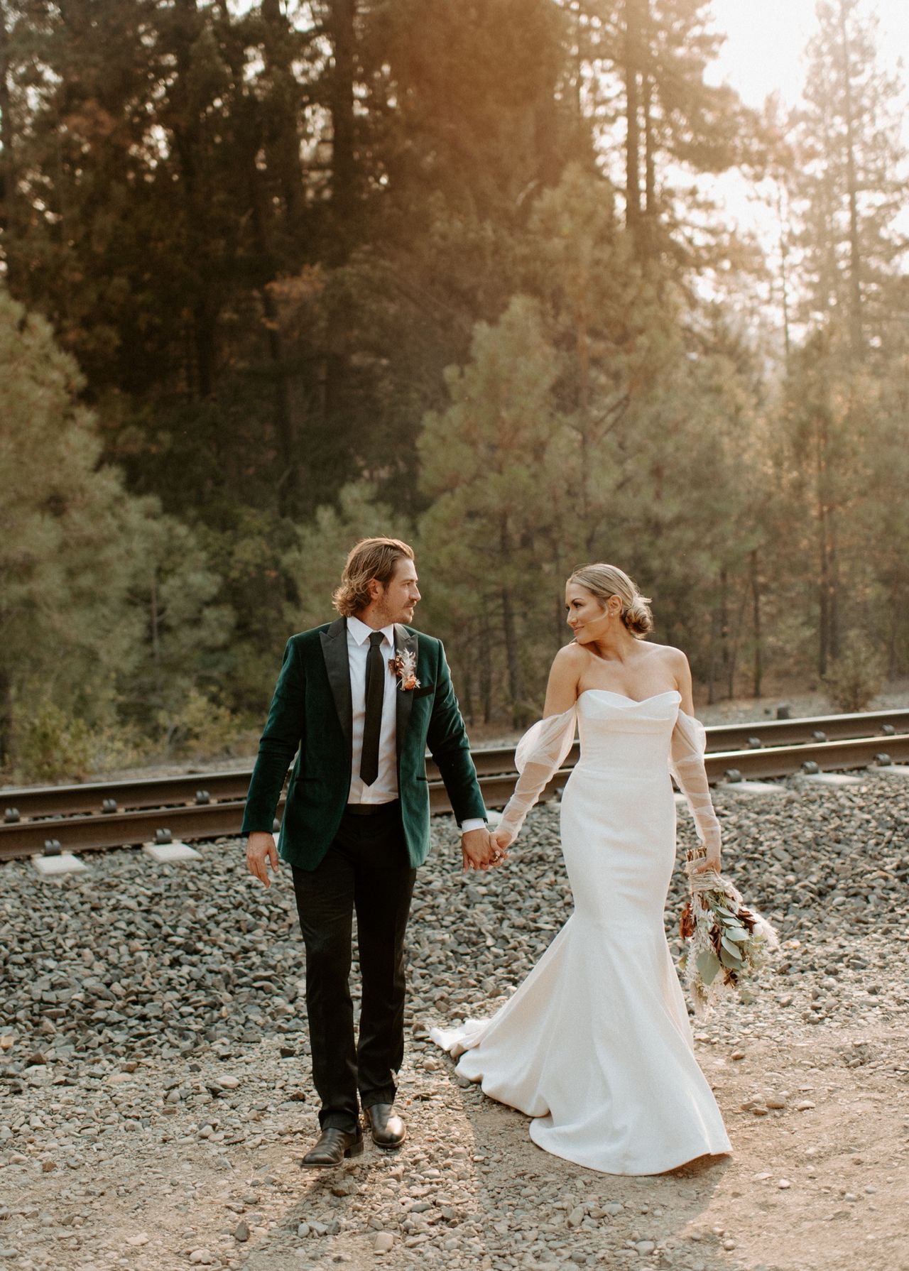 Joan and Kyle walk hand in hand beside the tracks in golden light