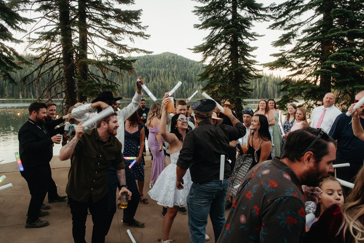 Kaitlyn laughs in white sunglasses as guests swing foam batons over her head by the lake