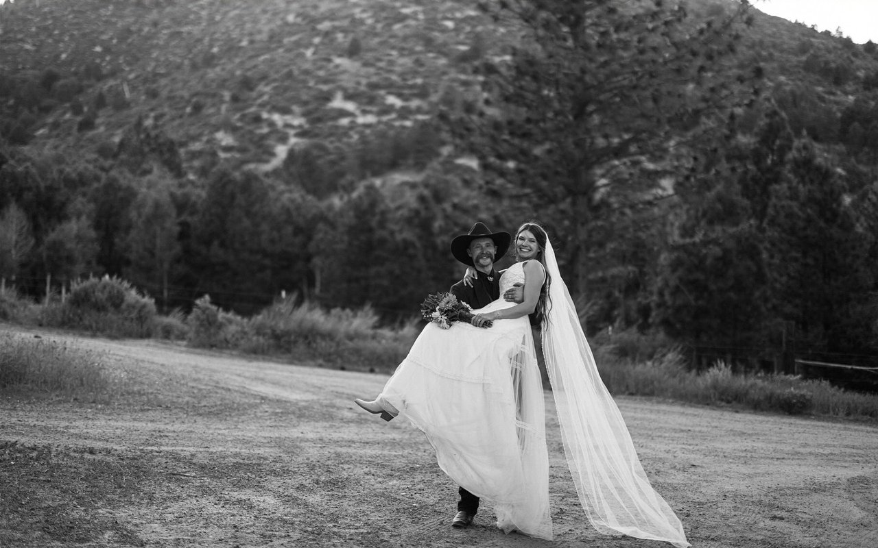 Jakob carries Kerstyn down the ranch road, her veil trailing, in black and white