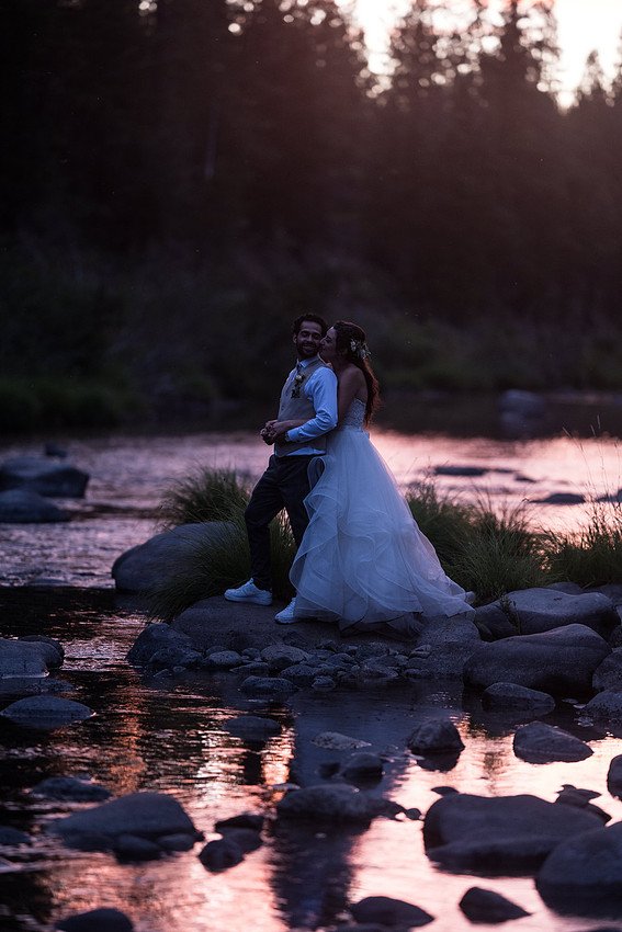 Shannon and Ryan lean together on the river rocks as the pink sky reflects in the water